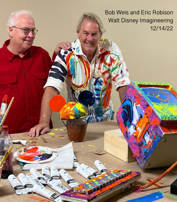 Two men with colorful Disney-themed paintings and supplies on table.