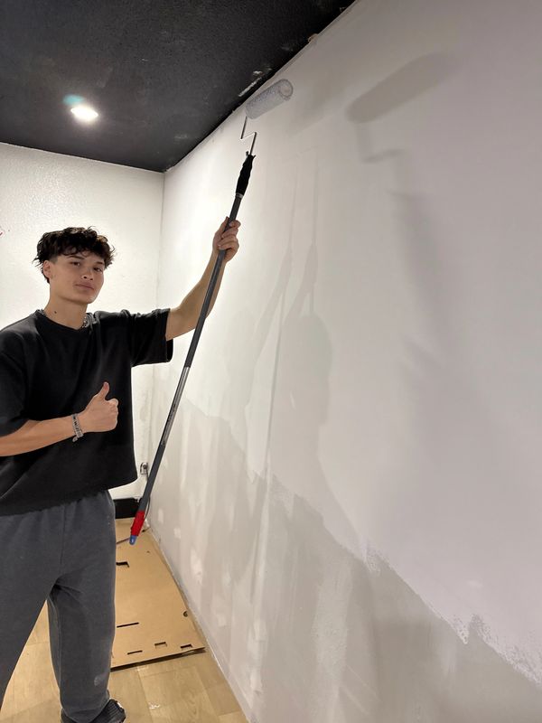 Young man painting a wall white with a roller and giving a thumbs up.