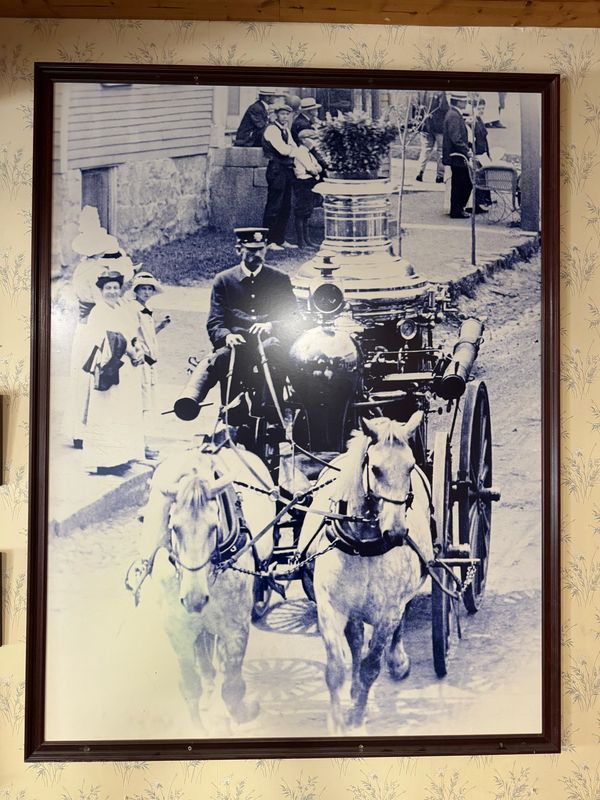 Vintage black and white photo of a horse-drawn fire engine with people in early 1900s attire.