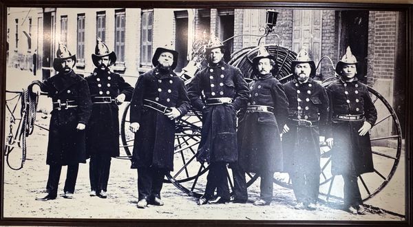 Vintage photo of seven firefighters in uniform standing in front of an old fire hose cart.