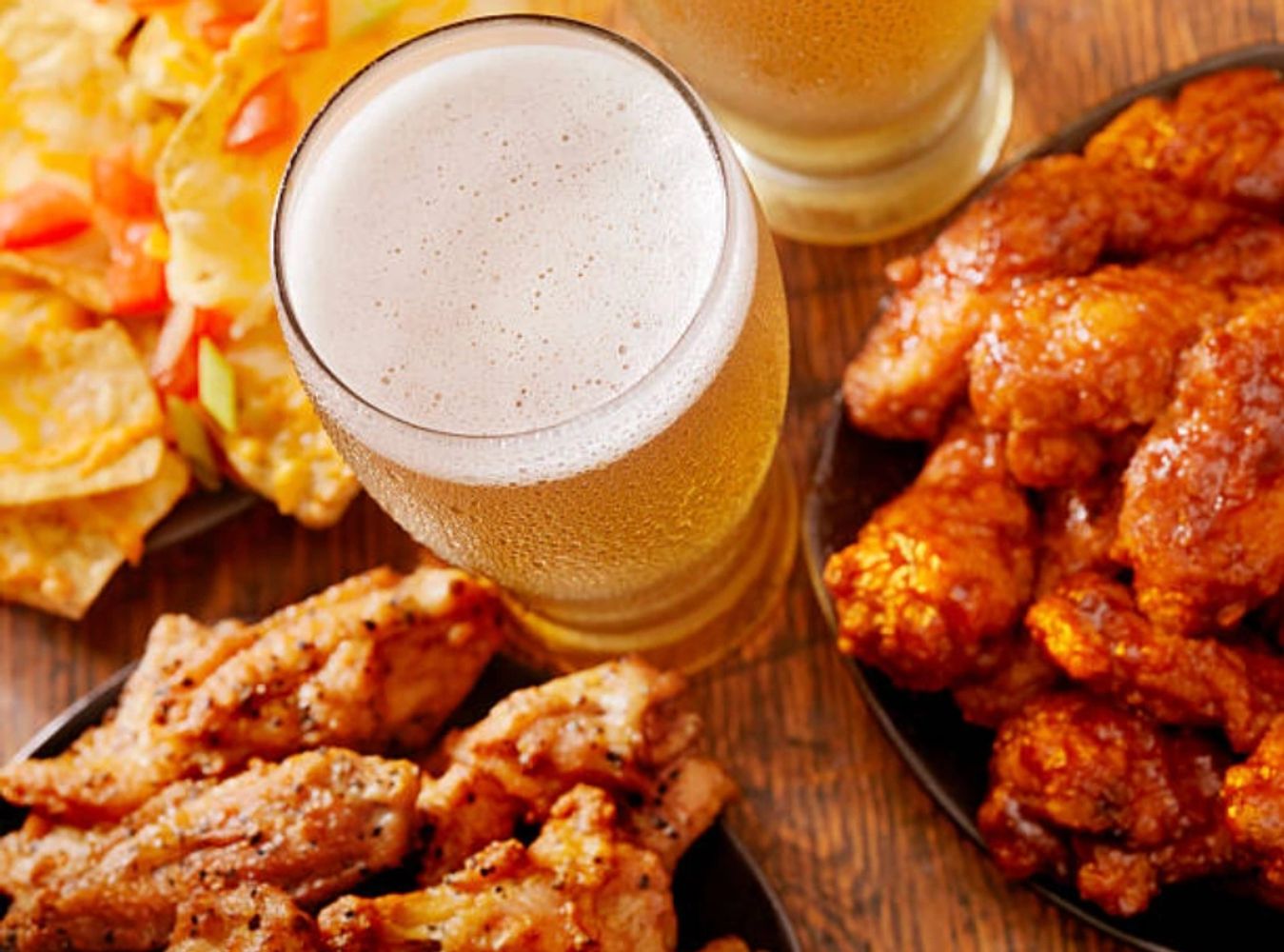 Beer glasses with crispy chicken wings and nachos on a wooden table.