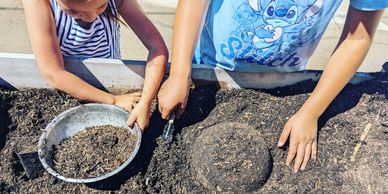 Two children playing with soil in a garden bed, making shapes with their hands and tools.