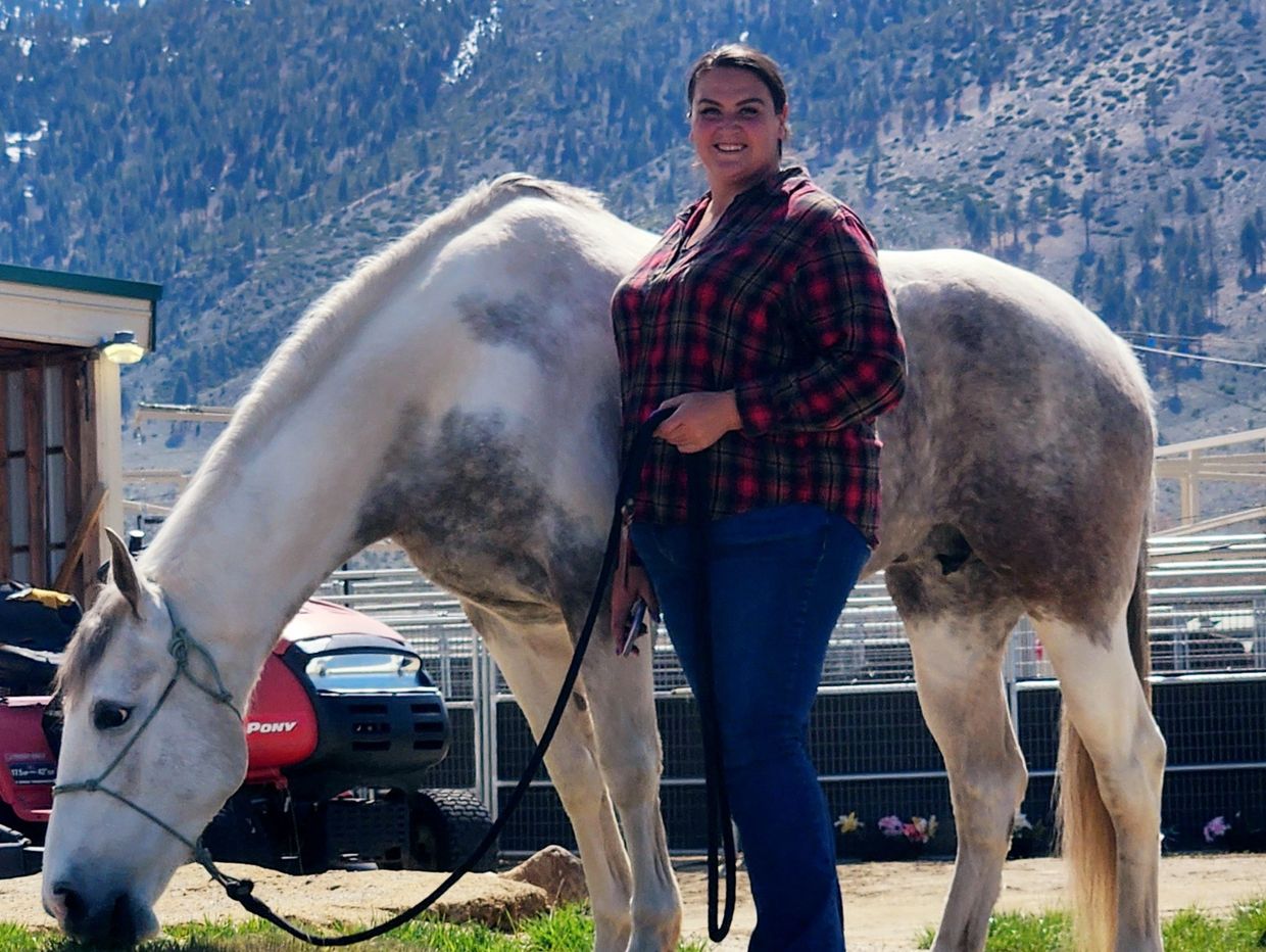 A woman in plaid stands beside a grazing horse with snowy mountains in the background.