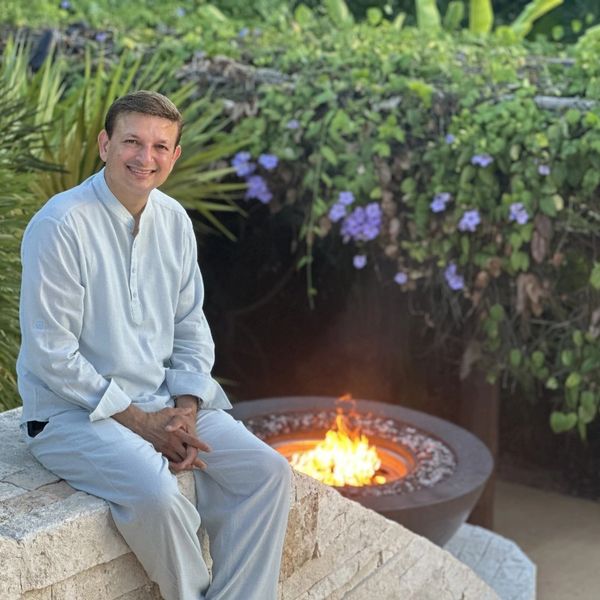 Man in white outfit sitting near an outdoor fire pit surrounded by greenery.