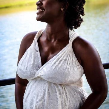 Pregnant woman in white dress smiling by water.