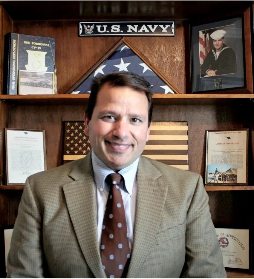 Man in a suit with U.S. Navy memorabilia behind him.