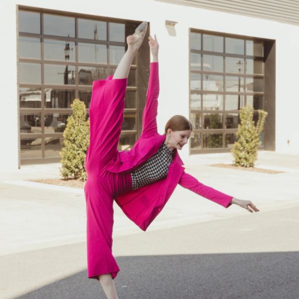 A woman in a bright pink suit performs a high leg stretch outdoors.