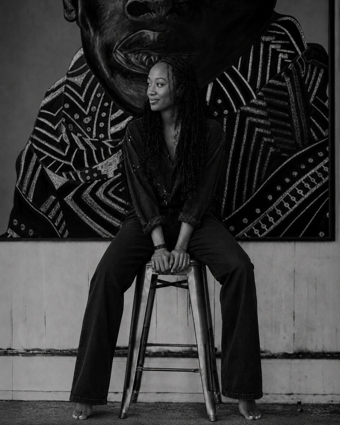 Woman sitting barefoot on a stool in front of a patterned mural.