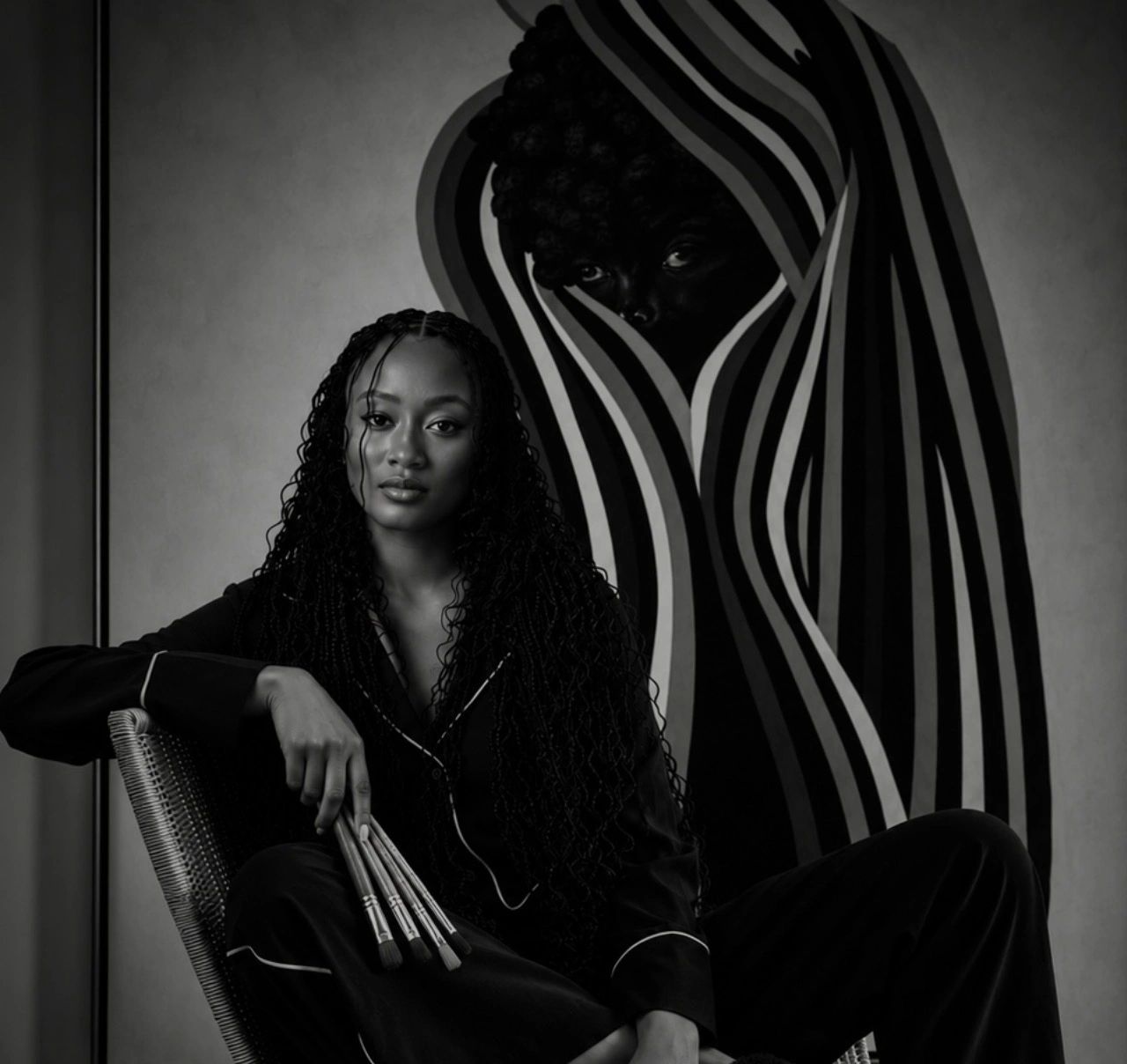 Woman with paintbrushes seated before an abstract striped artwork.