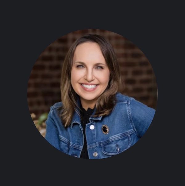 Smiling woman wearing a denim jacket against a blurred brick background.