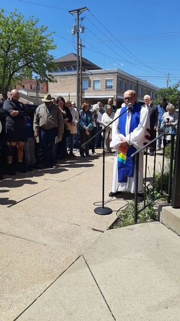 Clergy member addressing a gathered crowd outdoors on a sunny day.
