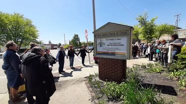 People gathered outside a church holding hands in a circle on a sunny day.