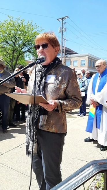 A woman speaks at an outdoor event with a microphone and clipboard.