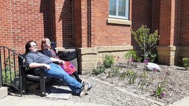 Two people relax on a bench outside a brick building on a sunny day.