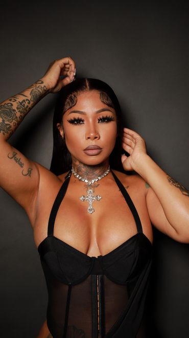 Confident woman in black corset with bold makeup and tattoos, posing against a dark background.