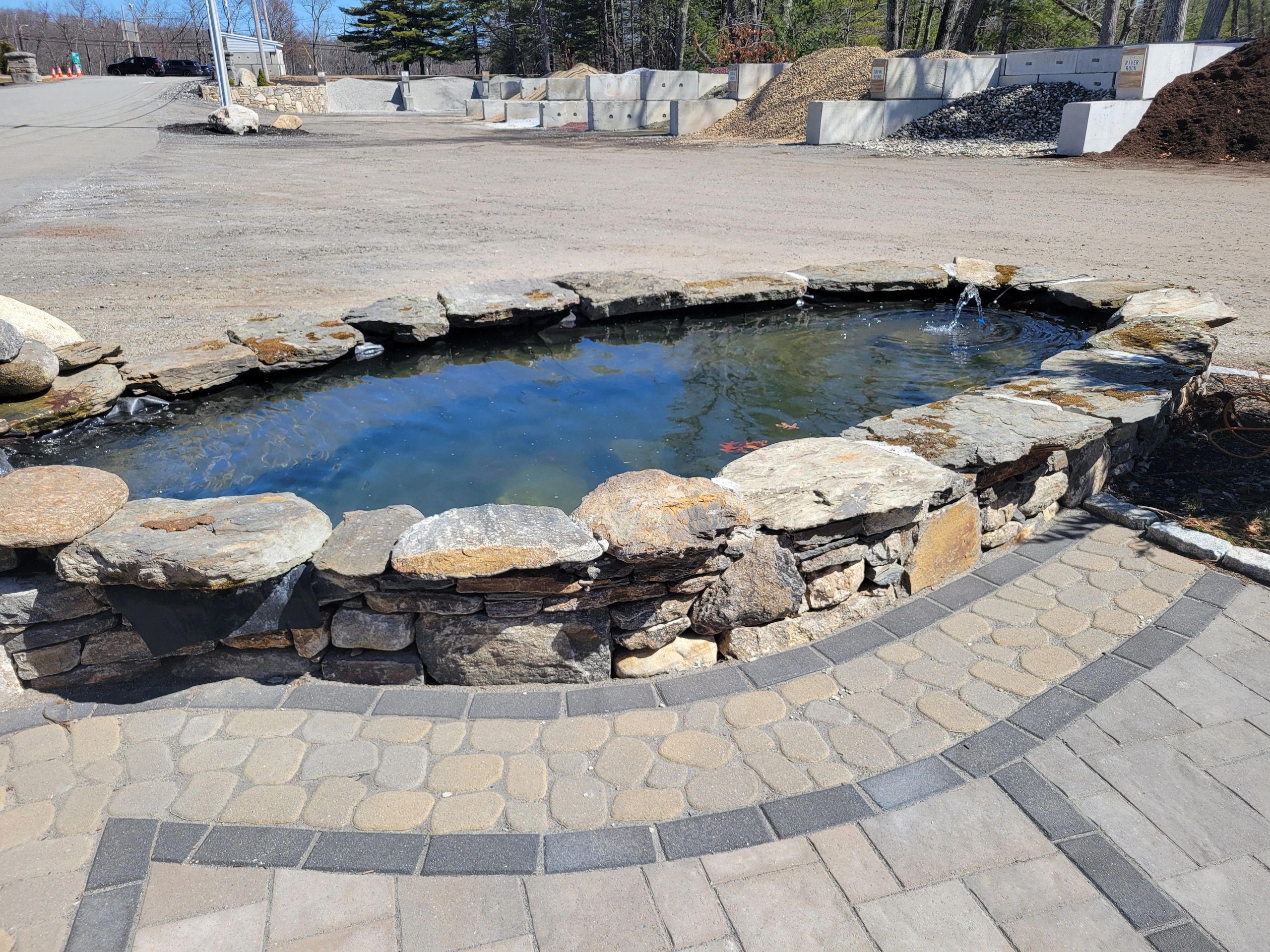 A small stone pond with a water fountain on a paved area outdoors.