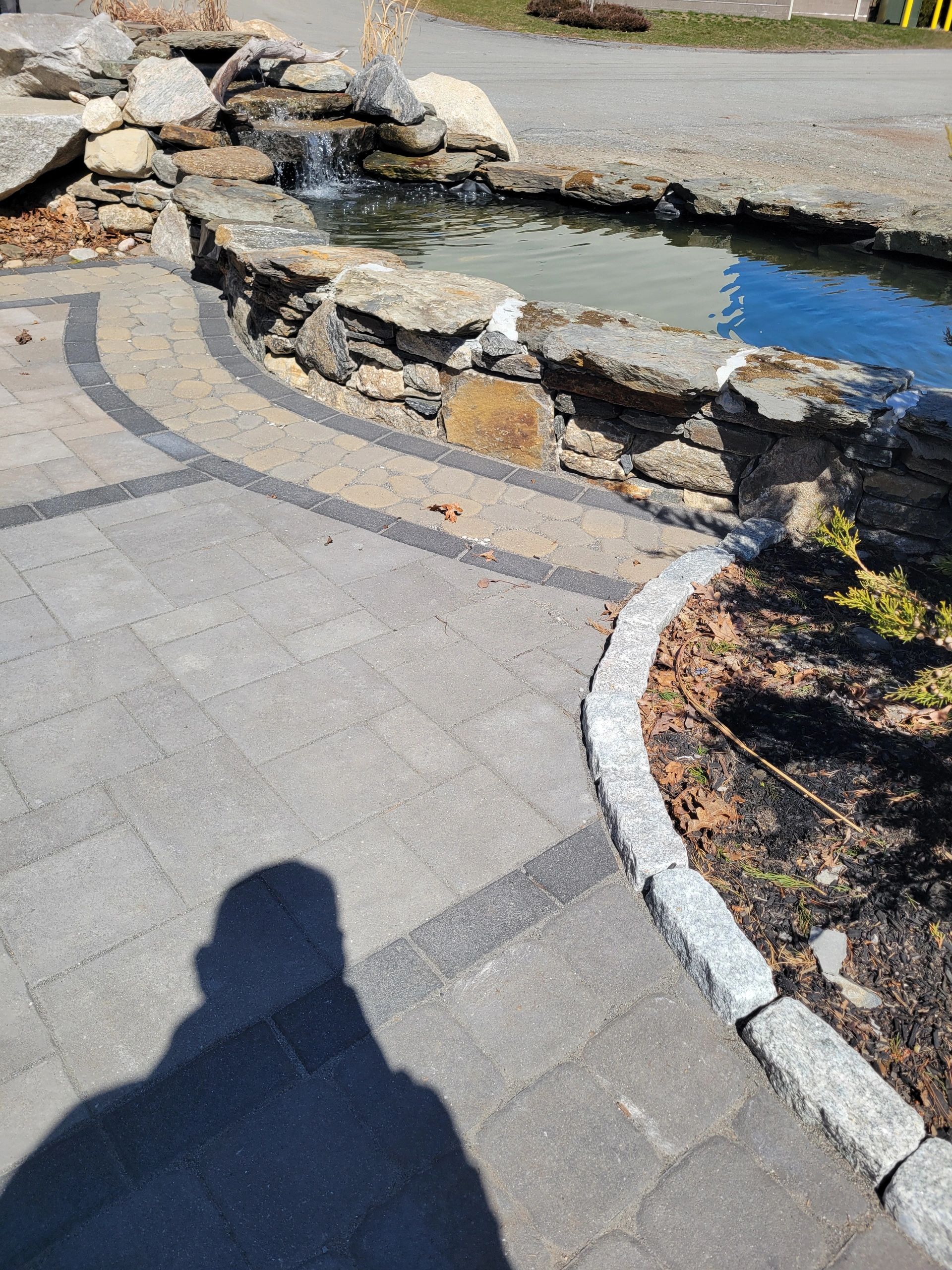 Stone pond with a small waterfall beside a paved walkway.