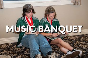 Two students wearing medals sit on the floor, writing in notebooks at a music banquet.