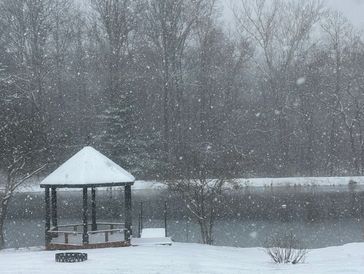 Snowfall covers a lakeside gazebo and snowy ground in a quiet winter scene.