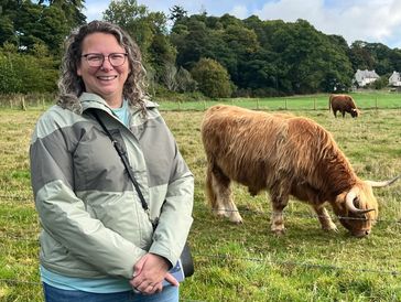 Woman smiling in a field with a Highland cow grazing nearby.