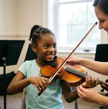 A young girl smiling while learning violin with an instructor.