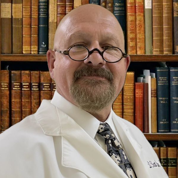 Smiling man in glasses and white coat stands before a bookshelf filled with old books.