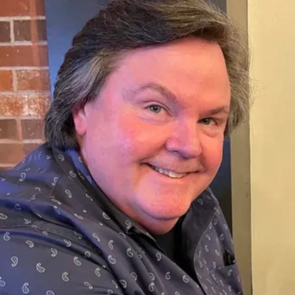 Smiling man with gray hair wearing a patterned blue shirt indoors.