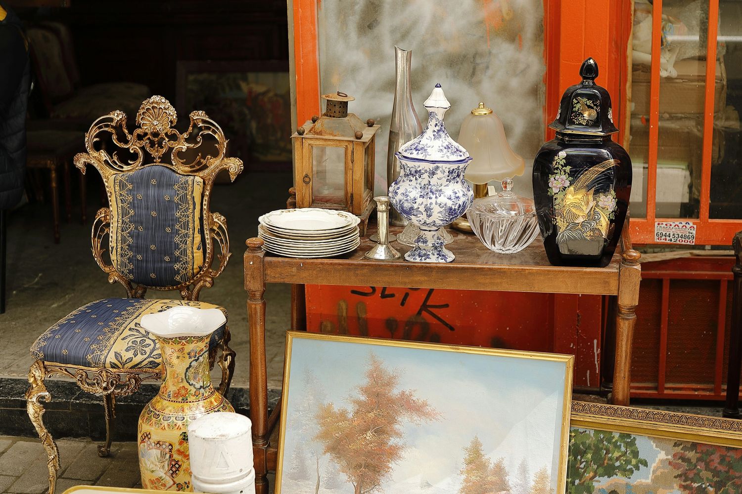 An antique shop display with ornate chair, vases, plates, and paintings.