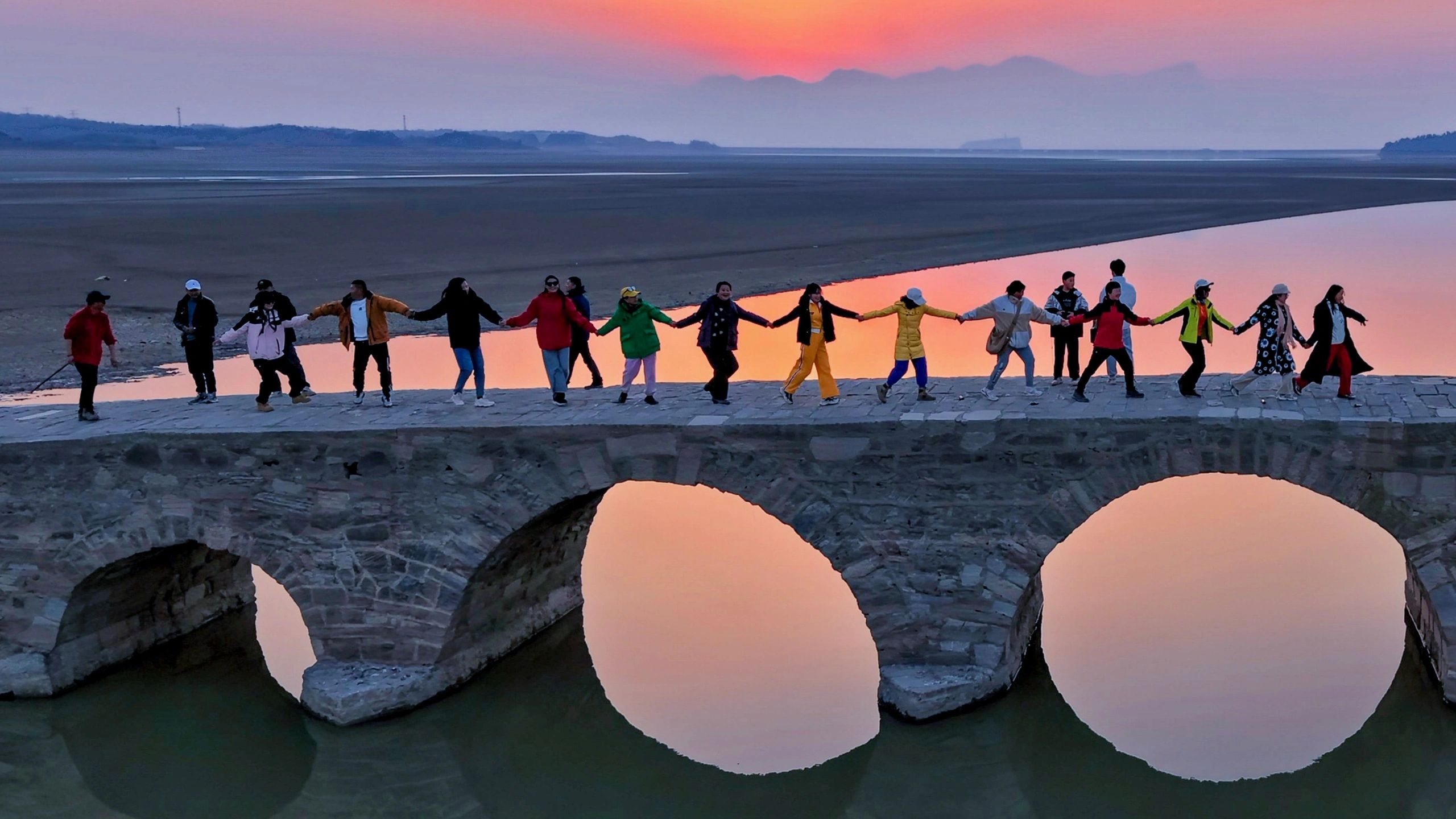 People holding hands on a stone bridge at sunset.