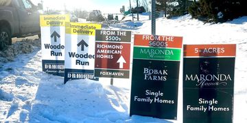 Real estate signs for homes and farms line a snowy roadside under a bright sun.