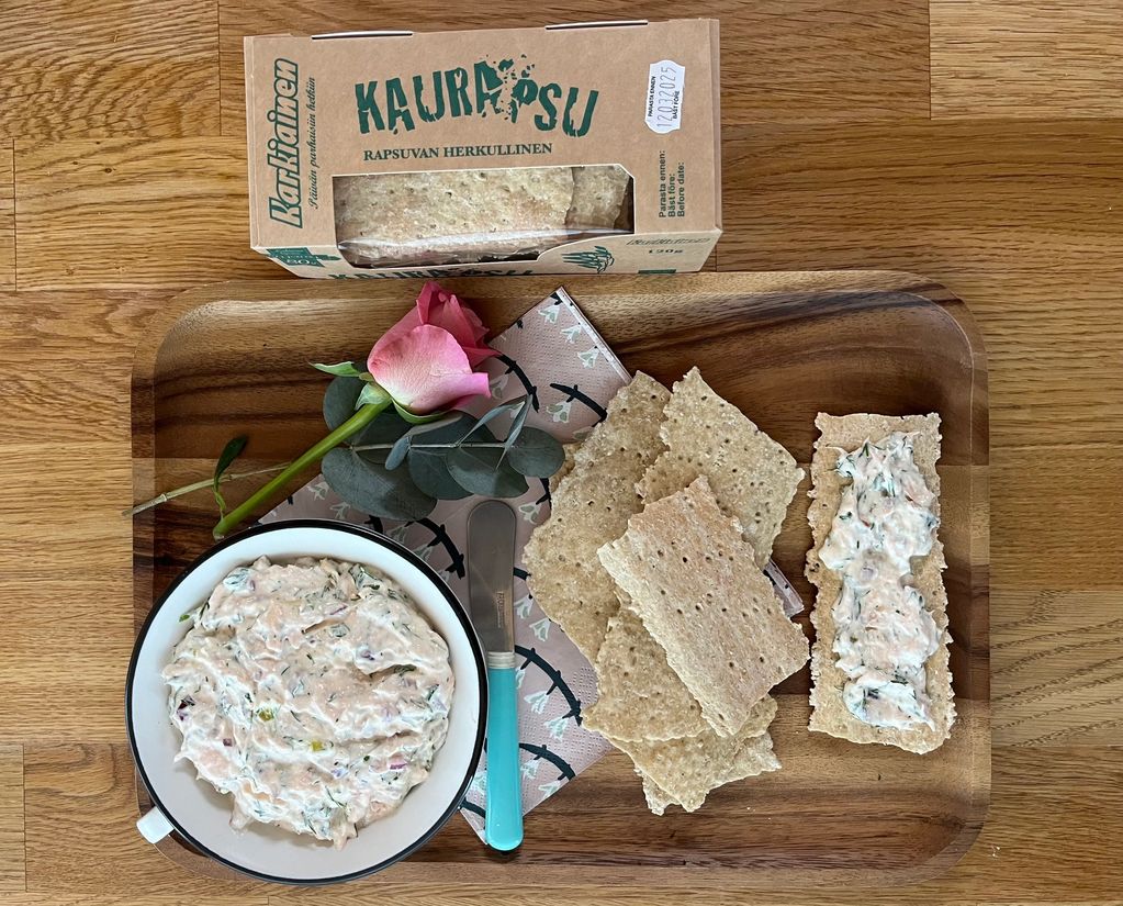 A wooden tray with oat crispbread, creamy spread, and a pink rose on a wooden table.