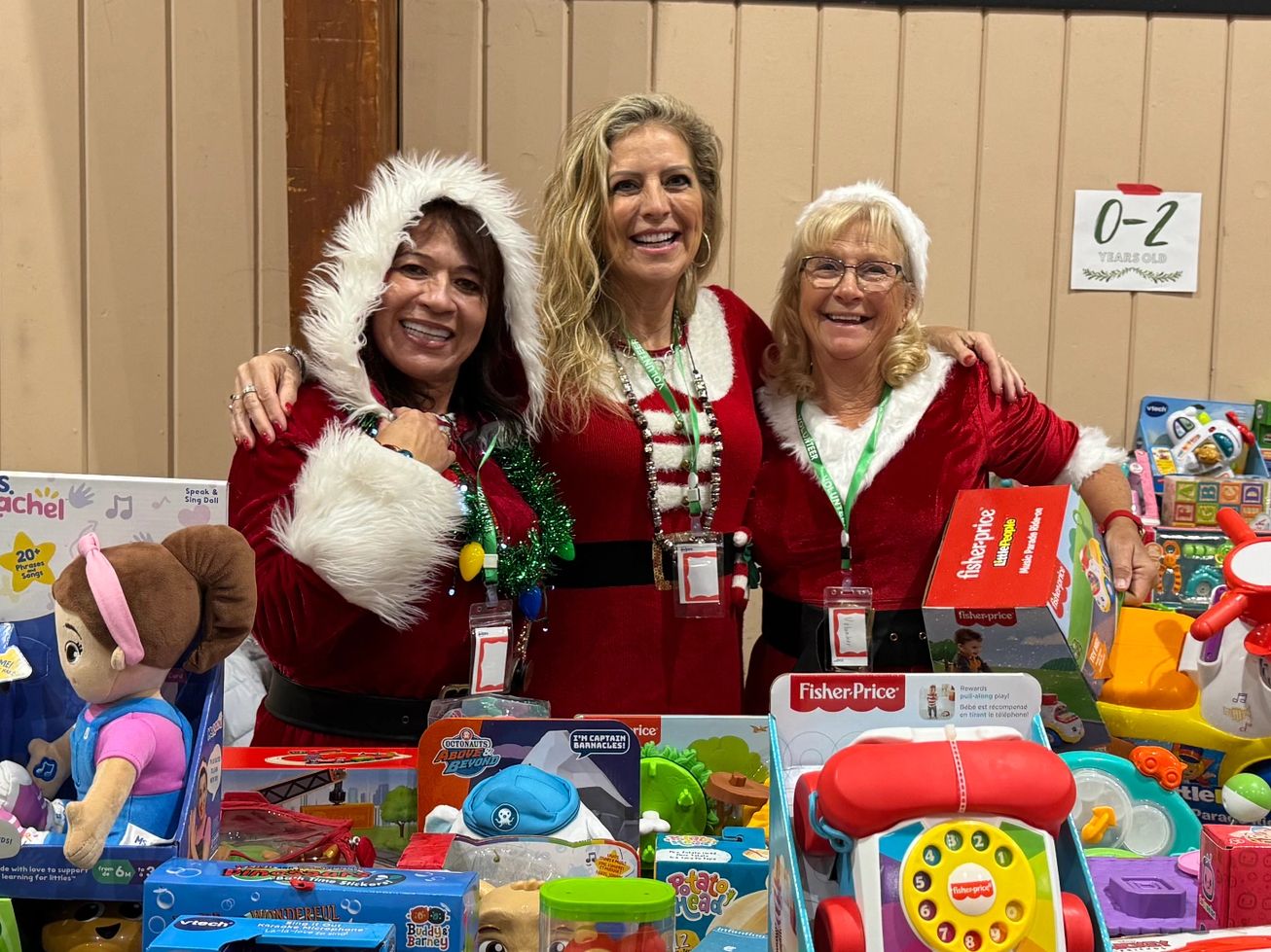 Three women dressed in festive red outfits smiling behind a table full of toys.