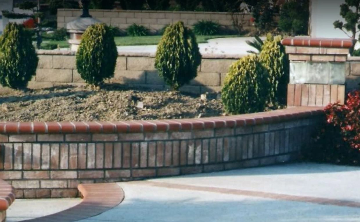 Curved brick retaining wall with small green shrubs and a concrete driveway.
