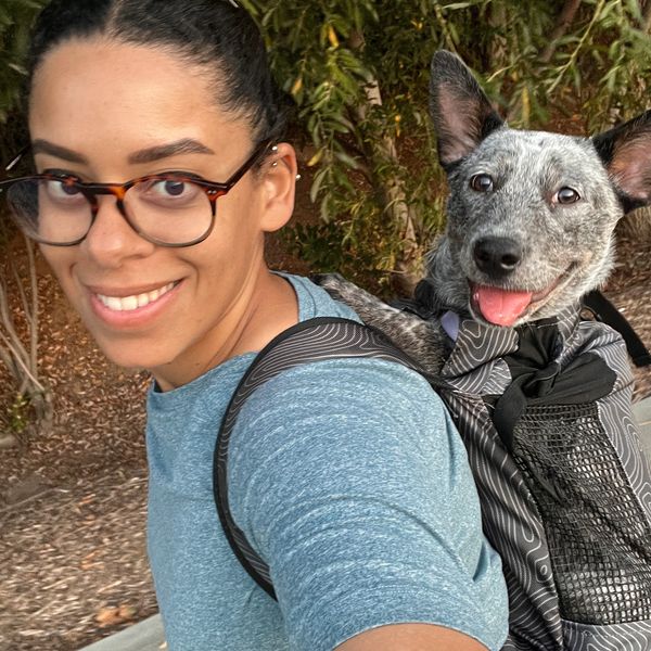Smiling woman with glasses carrying a happy dog in a backpack outdoors.