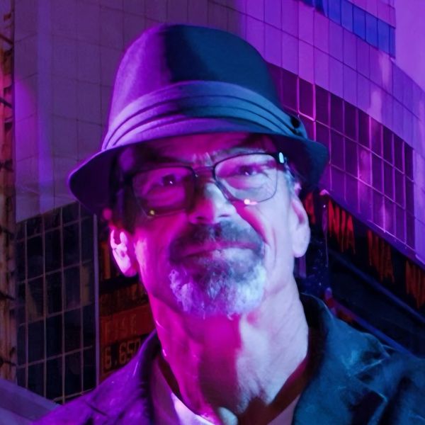 Man with glasses and hat illuminated by purple light in urban setting.