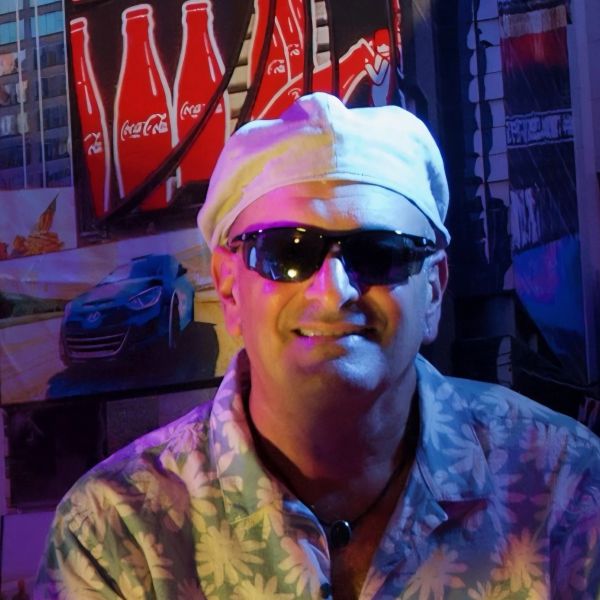 Man wearing a white cap and sunglasses in a colorful, neon-lit setting.
