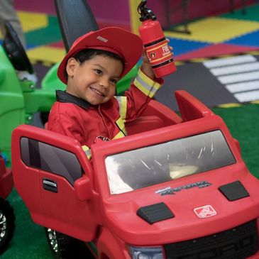 Smiling child dressed as firefighter in a red toy car holding a fire extinguisher.