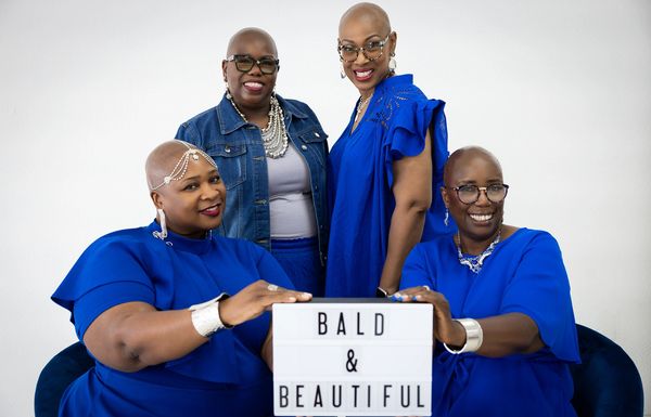 Four stylish bald women in blue outfits hold a sign reading 'Bald &amp; Beautiful.'