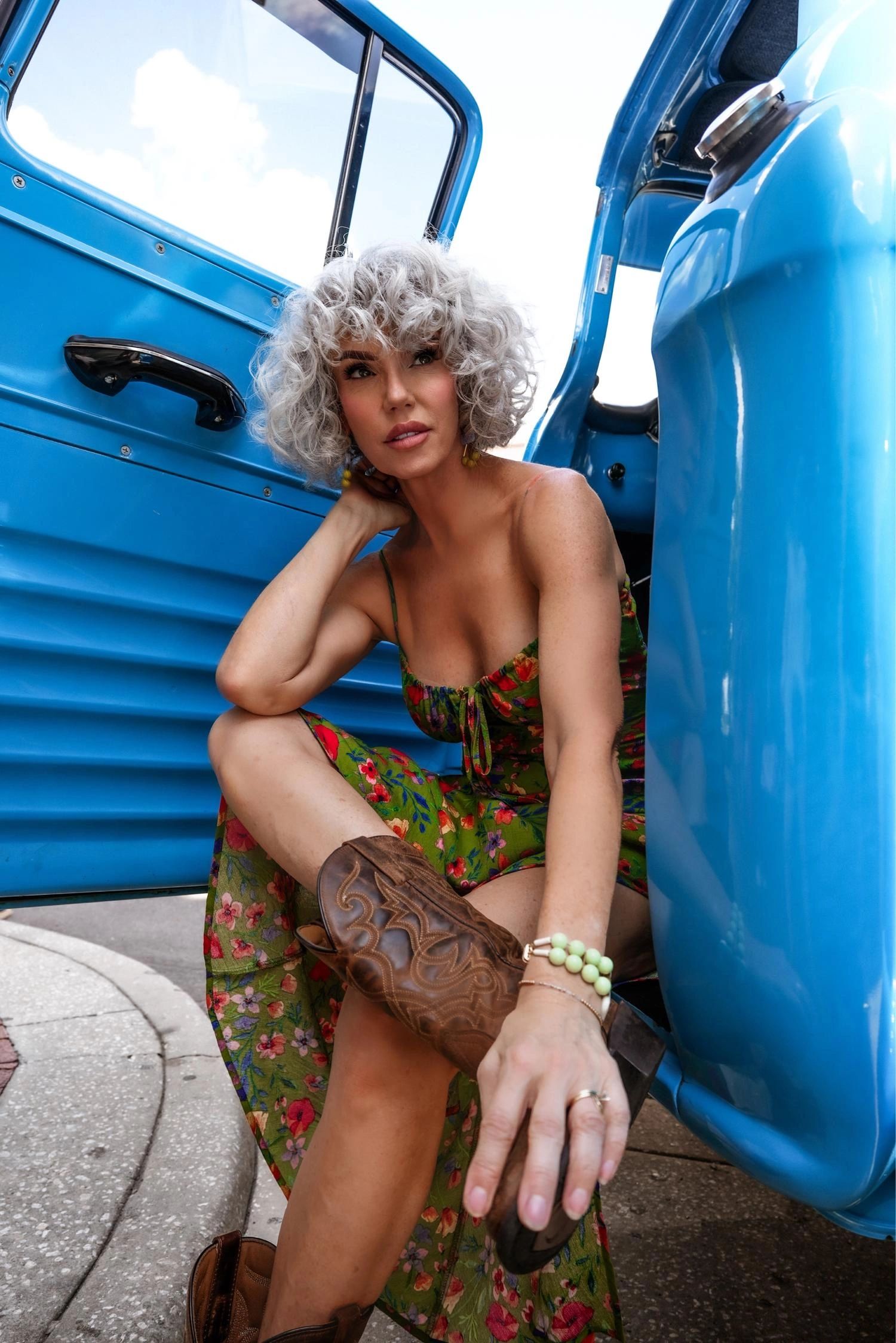 Woman with curly gray hair sitting by an open blue car door, wearing a floral dress and brown boots.