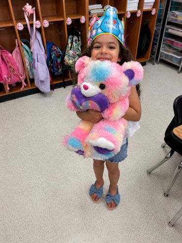 Smiling girl wearing a birthday crown hugging a colorful teddy bear in a classroom.