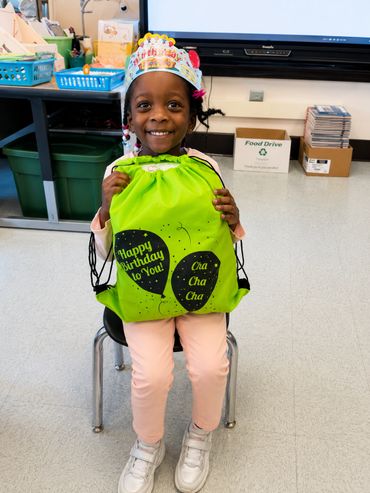 A smiling girl wearing a birthday crown holds a green birthday gift bag in a classroom.