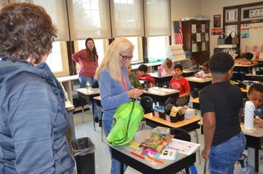 Teacher organizing supplies in a busy classroom with students and another adult present.