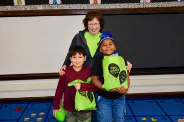 Teacher with two young students holding green bags in a classroom.