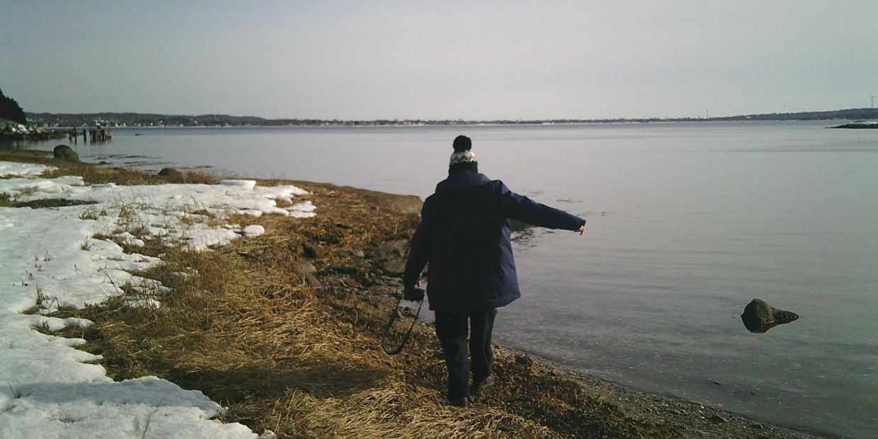 Person walking along a snowy and grassy lakeshore on a calm day.