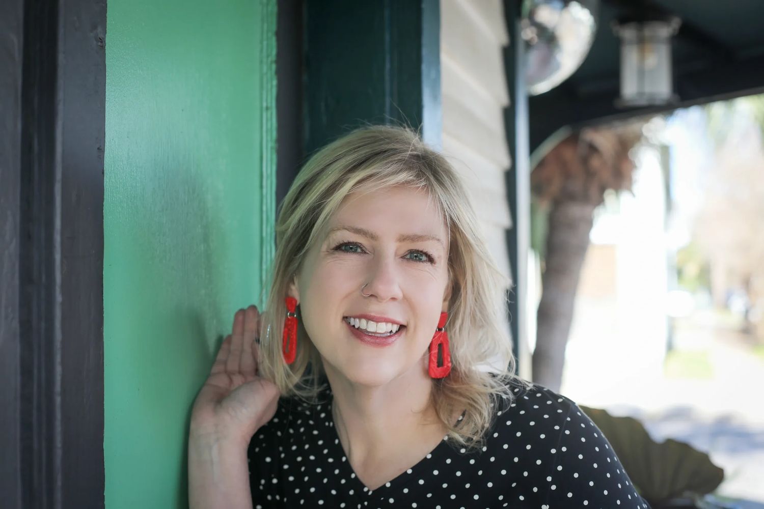 Smiling woman with blonde hair and red earrings leans against a green door.