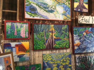 Colorful landscape paintings displayed on a rustic wooden wall with string lights.