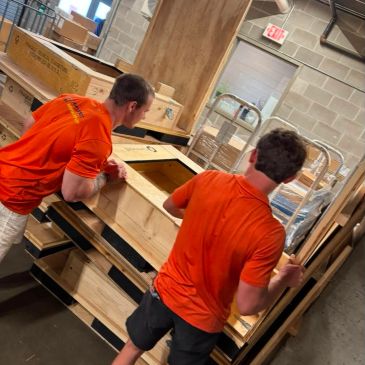 Two men in orange shirts moving wooden crates in a warehouse.