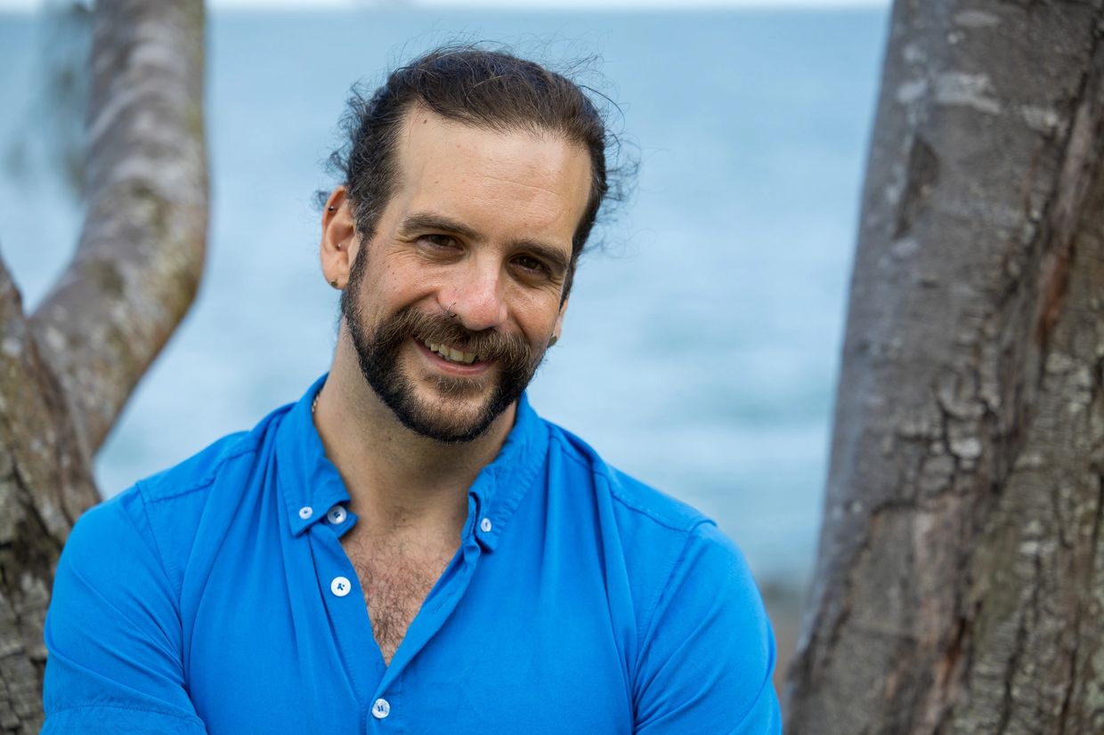 Smiling man in a blue shirt outdoors near trees and water.