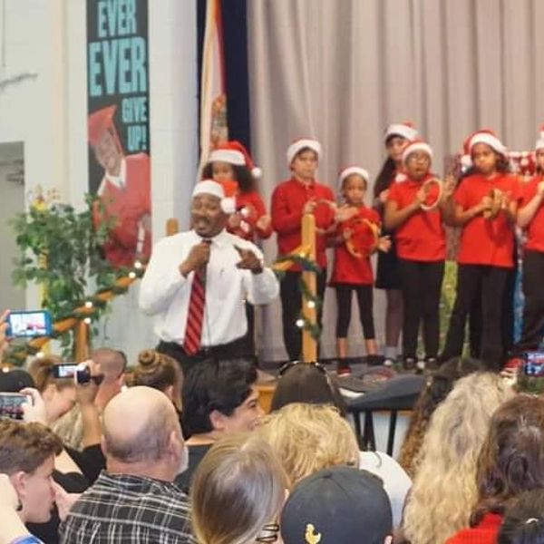 Children in Santa hats performing on stage with a speaker addressing the audience.