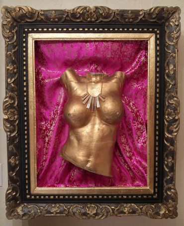 Golden female torso sculpture framed with ornate border and pink fabric.